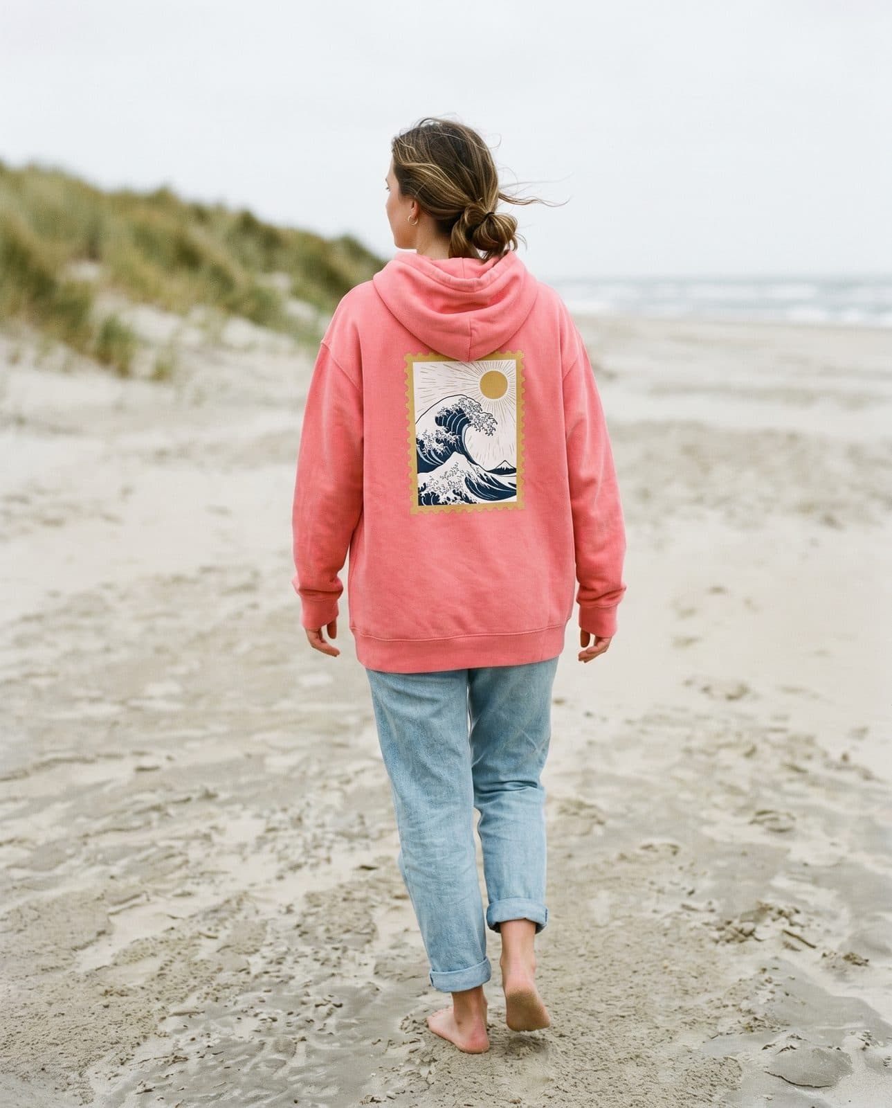 Frau im korallenfarbenen SurferAI-Hoodie „Die Welle“ am Strand von Langeoog